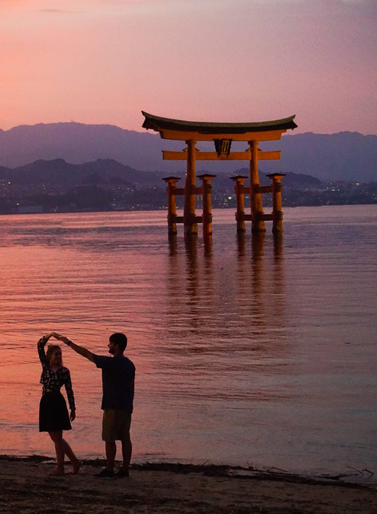 Woman being twirled by a man (dancing) in front of a orange torii gate (shrine) at sunset. A beautiful view on a day trip to Miyajima Island.