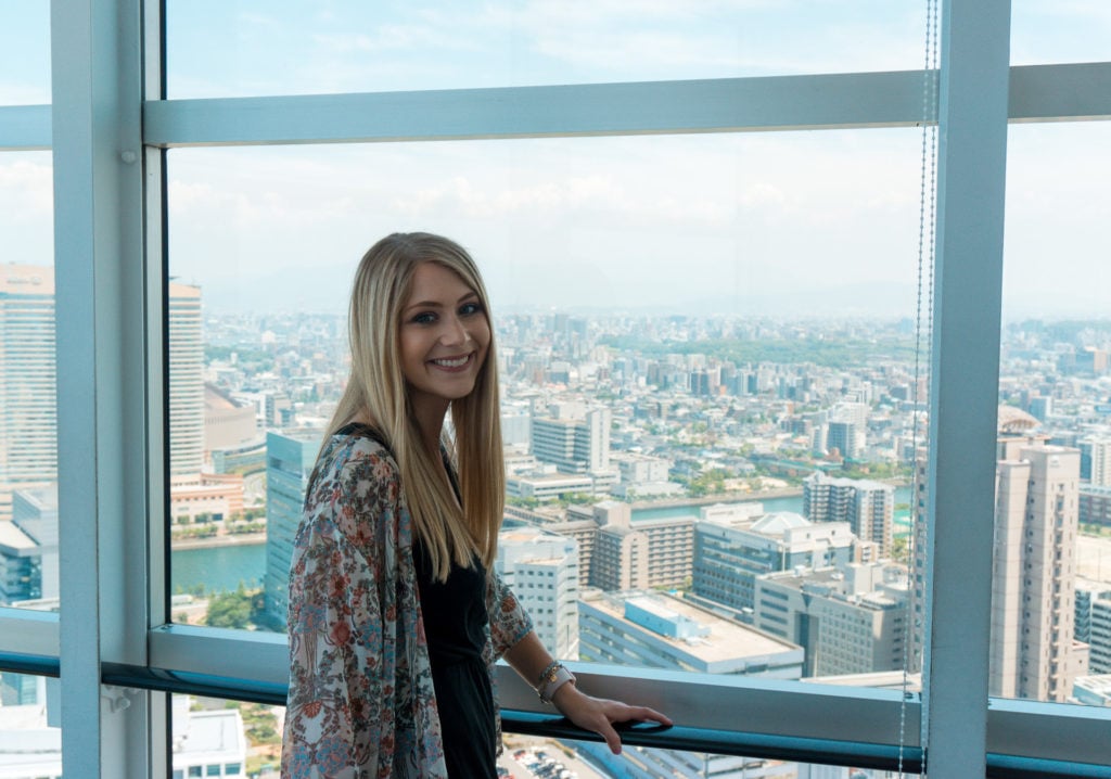 Woman standing by a window with a city skyline in the background (Fukuoka itinerary)