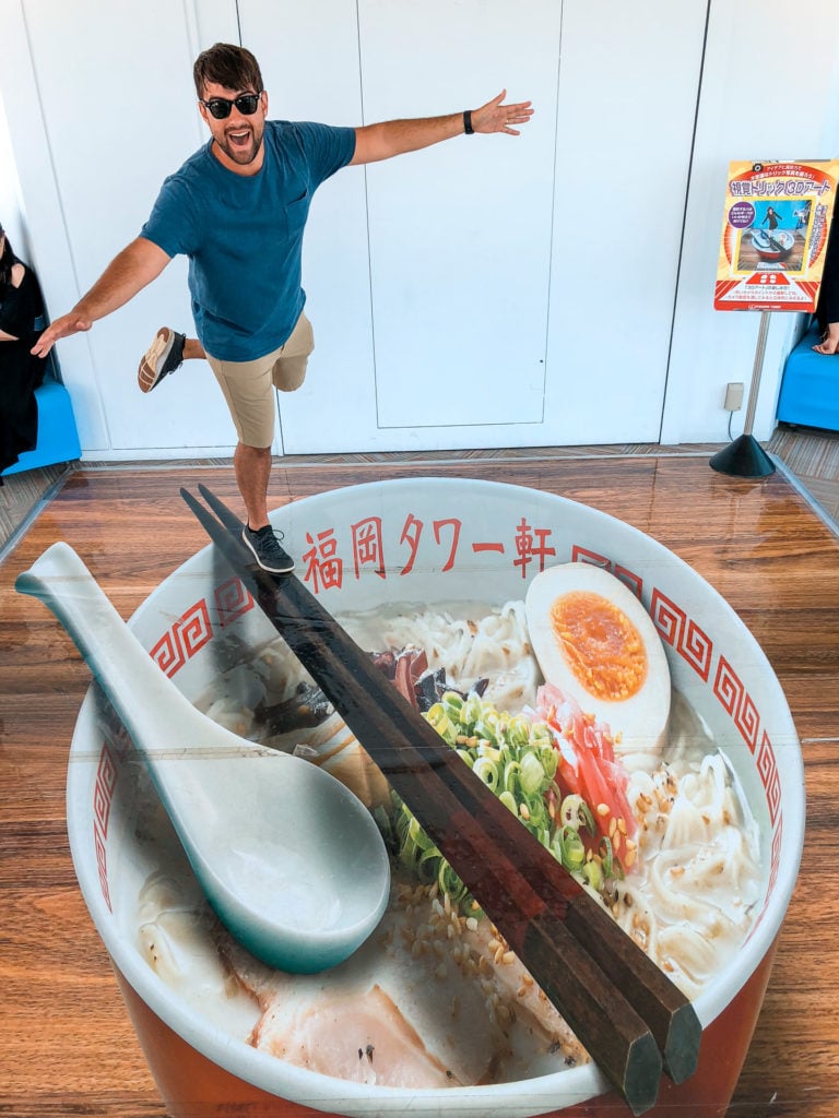 Man standing over a large display of ramen and pretending to balance on faux chopsticks at Fukuoka Tower.