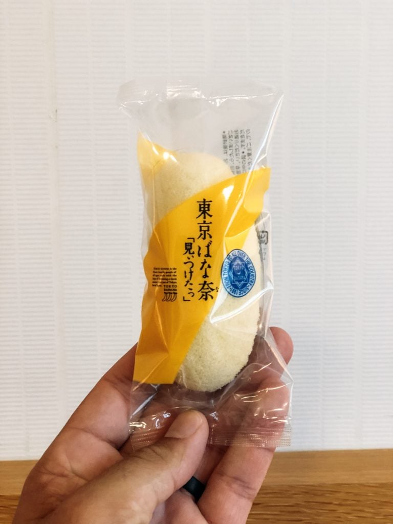 Hand holding a packaged light yellow banana shaped cake dessert (Tokyo banana)