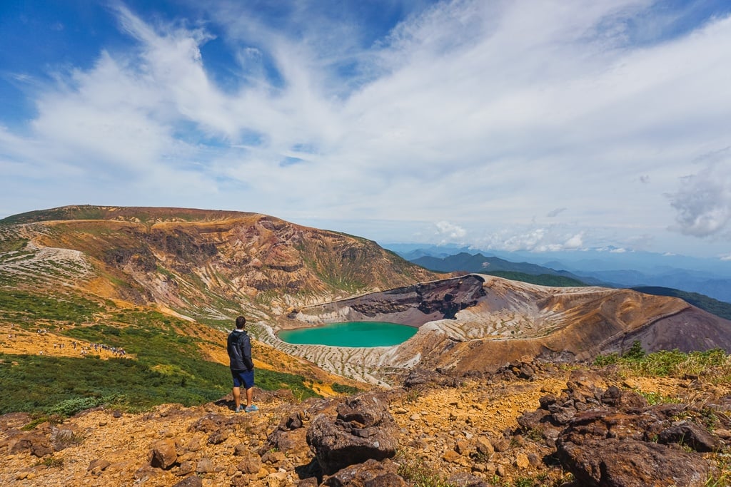 Man with dark hair dressed in a black jacket and black shorts standing in front of Okama Crater from Mt. Zao in Japan. 